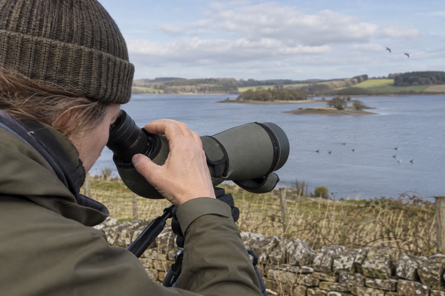 Observer turning a spotting-scope zoom eyepiece beside a reservoir
